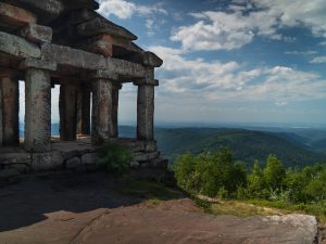 Ruins of a stone structure with pillars overlooking a lush green valley and distant hills under a partly cloudy sky.
