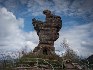 A tall rock formation resembling a giant sculpture, surrounded by a green railing and trees, under a cloudy sky.