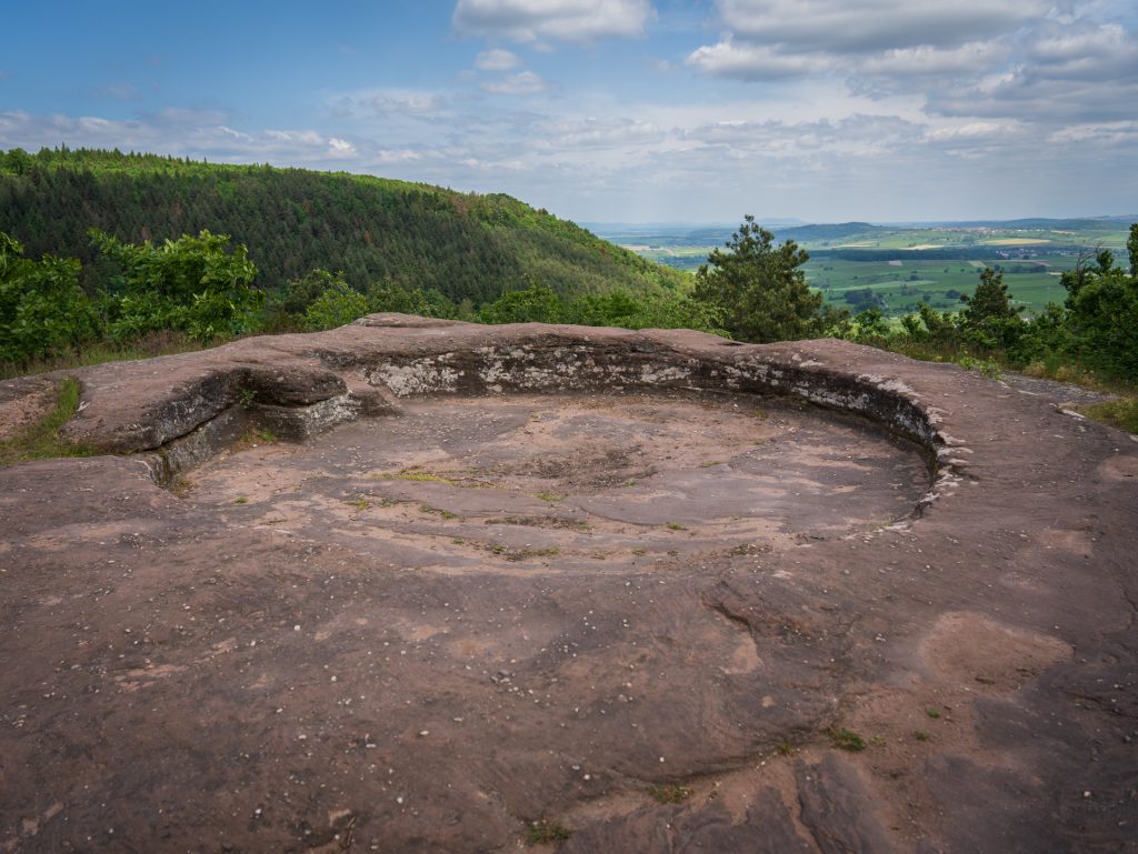 A circular stone platform with a rocky texture, surrounded by a lush green landscape and distant hills under a cloudy sky.