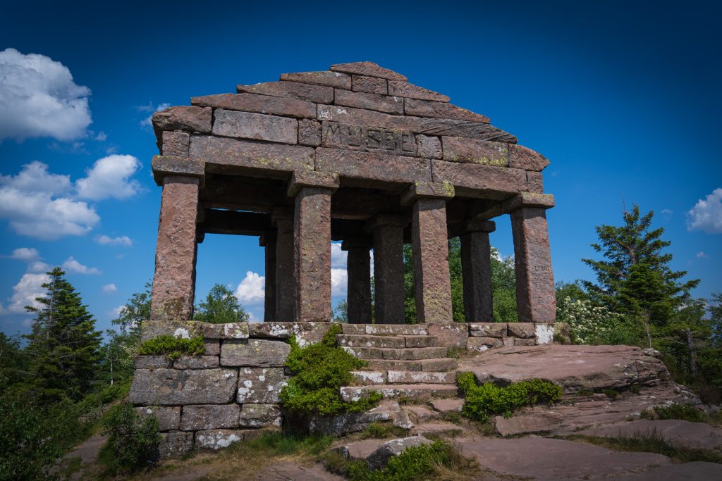 A stone structure with a triangular roof supported by four columns, surrounded by greenery and under a blue sky with clouds.