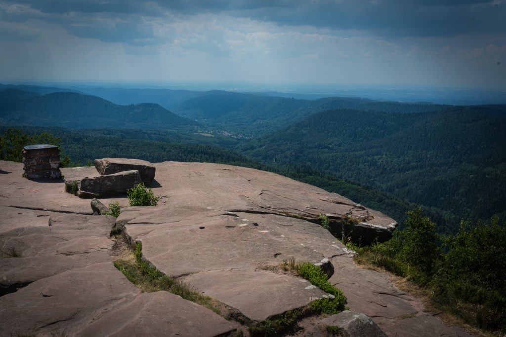A rocky overlook with a flat surface leads to a sprawling view of green hills and valleys under a cloudy sky.