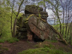 A large, mossy rock formation amidst lush green trees, featuring a cave-like opening on one side.