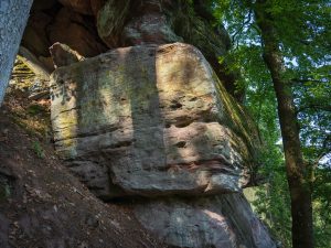 A large, moss-covered rock formation partially shaded by trees, with textures and colors indicating weathering and time.
