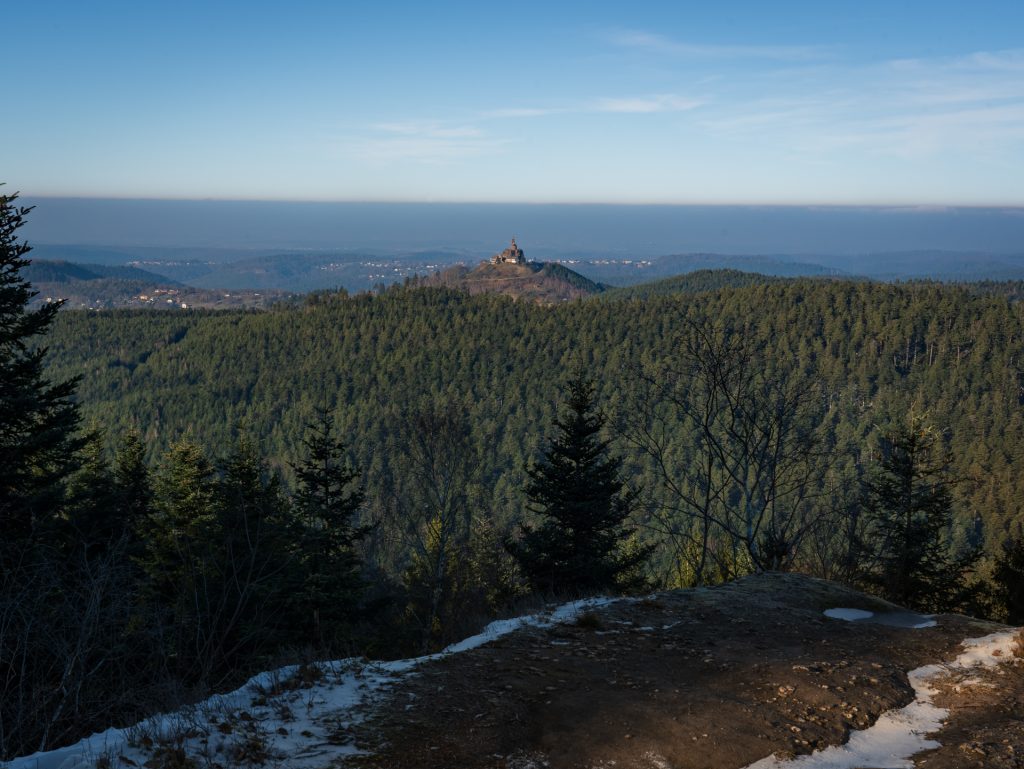 A panoramic view of a mountainous landscape with evergreen trees and a distant hilltop structure under a clear blue sky.