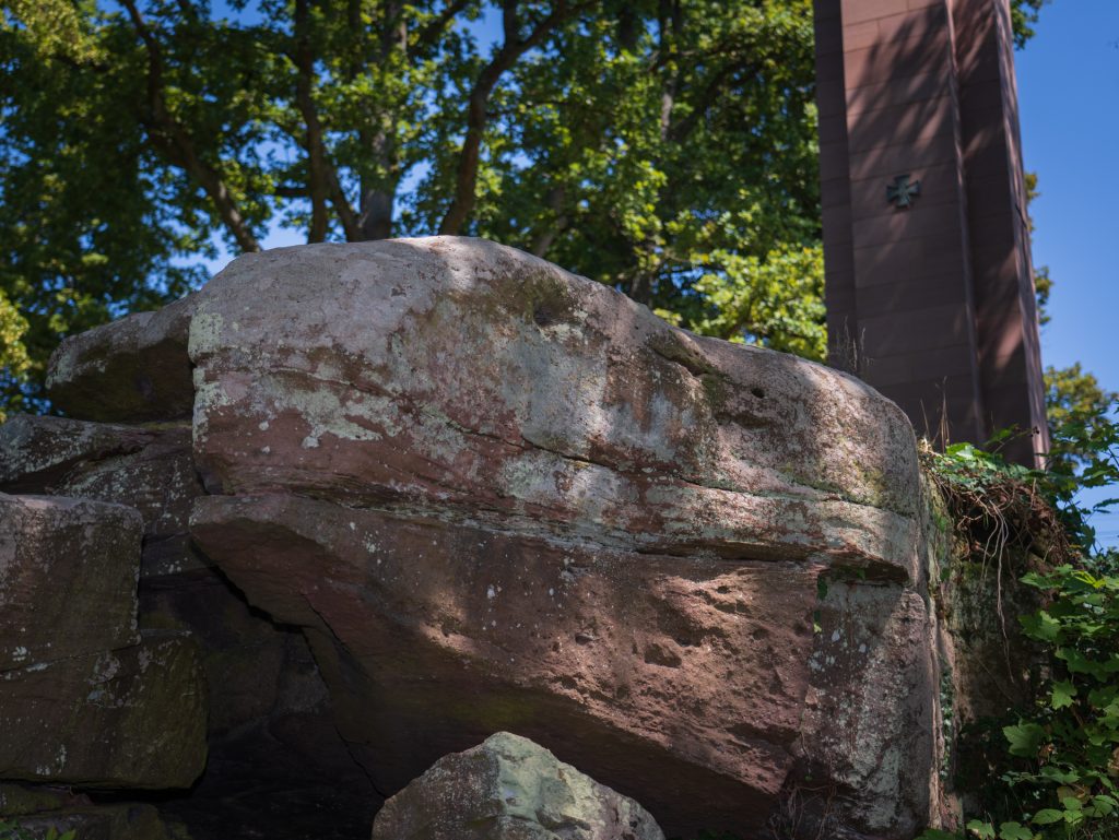 A large, weathered rock covered in green moss and lichen, with a tall structure and trees in the background.