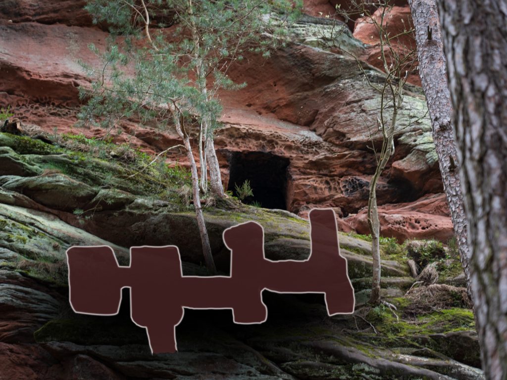 A moss-covered rock face with a dark cave entrance, framed by trees, against a backdrop of red sandstone cliffs.