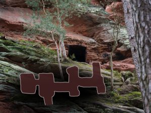 A moss-covered rock face with a dark cave entrance, framed by trees, against a backdrop of red sandstone cliffs.