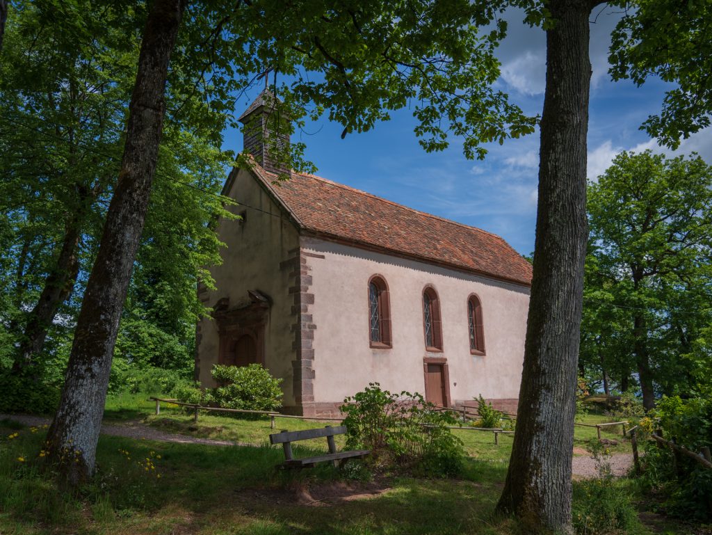 A quaint church with a simple facade and a red-tiled roof, surrounded by trees and a grassy area under a blue sky.