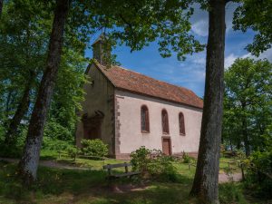 A quaint church with a simple facade and a red-tiled roof, surrounded by trees and a grassy area under a blue sky.