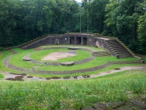 An ancient, circular amphitheater surrounded by lush green trees and slightly damp ground, showcasing tiered seating.