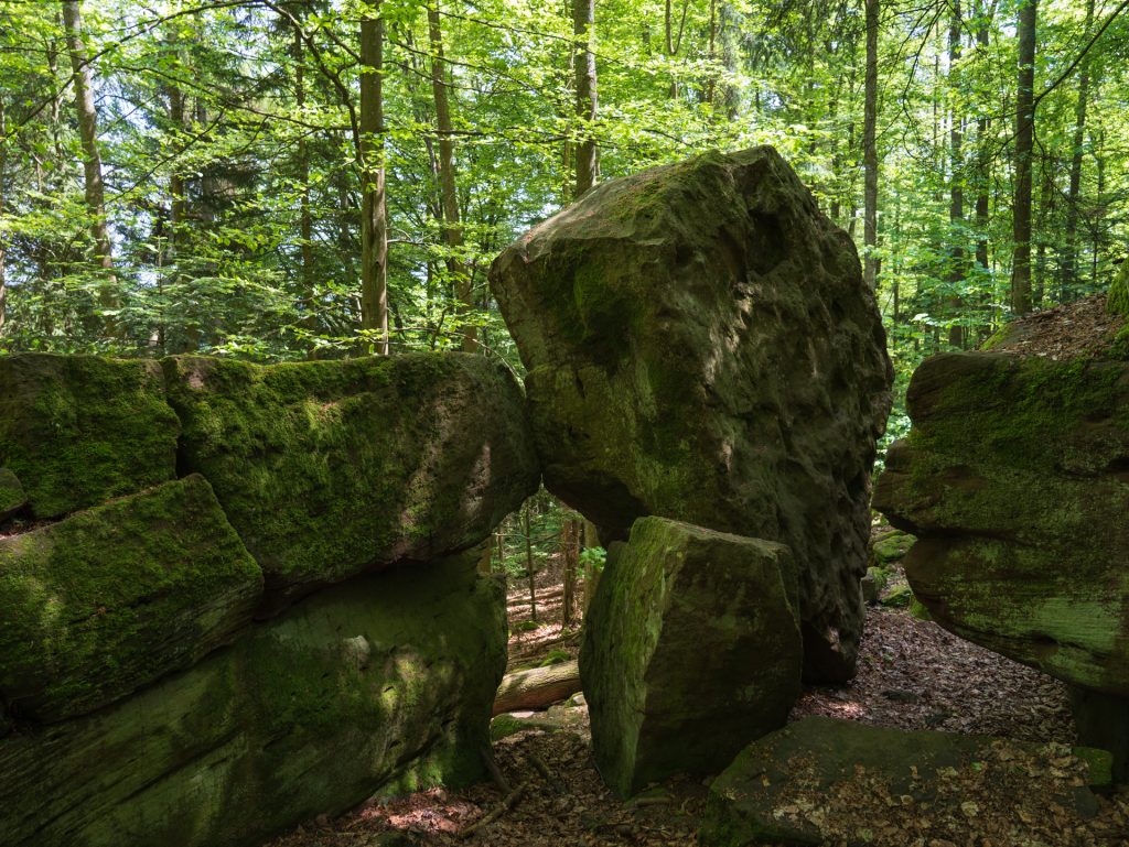 A large, moss-covered boulder is wedged between two stone formations in a lush green forest. Sunlight filters through the trees above.