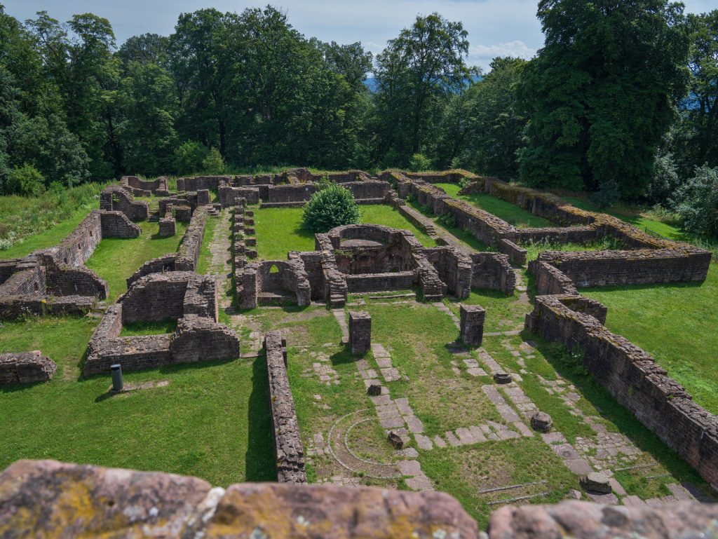 Ruins of an ancient stone structure surrounded by greenery, featuring stone walls and pathways in a rectangular layout.
