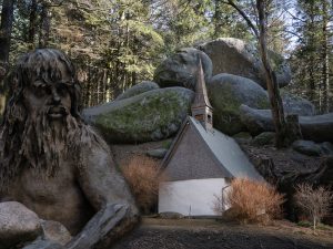A large stone statue of a bearded man sits beside a small, white chapel with a steep roof, surrounded by boulders and forest.