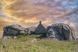 A rustic stone house nestled among large boulders, set against a colorful sky with clouds at sunset.