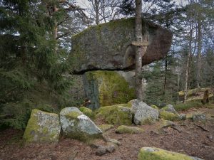 A large boulder rests atop a smaller moss-covered rock in a forested area, surrounded by trees and smaller stones.