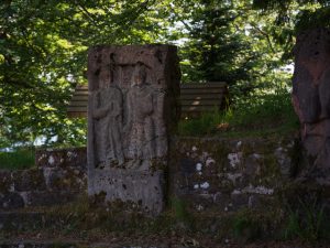 Weathered stone relief of two standing figures in a wooded, mossy outdoor setting